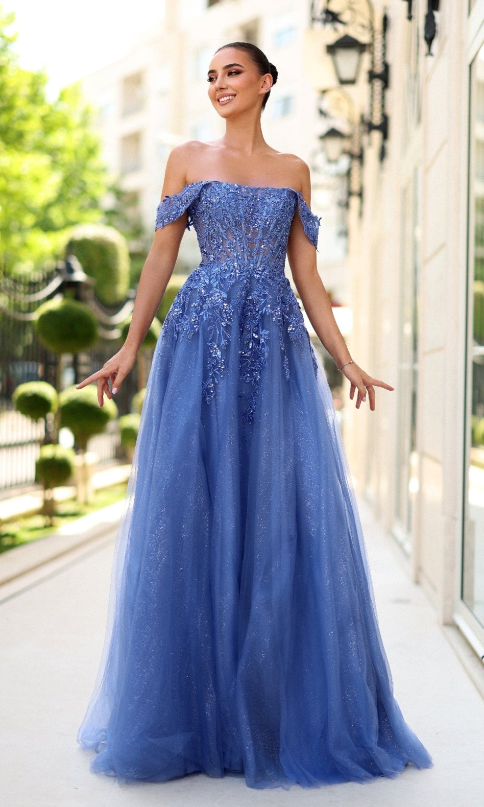 A woman stands outdoors in the Long Glitter Blue Prom Dress: Tina Holly TE232, featuring an off-the-shoulder corset bodice, sparkling floral embellishments, and a glitter tulle skirt, smiling with arms slightly raised.
